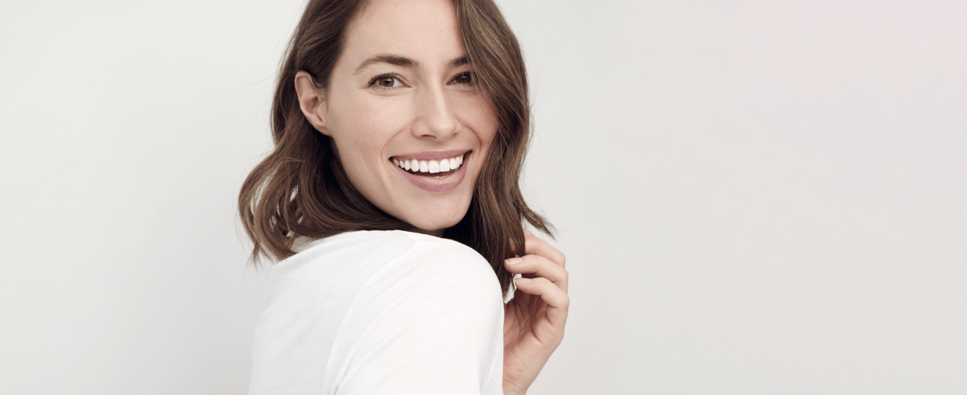 A woman with shoulder-length brown hair smiles brightly, looking over her shoulder. She wears a white shirt and stands against a plain, light background, radiating confidence often seen in the clients of a plastic surgery specialist.