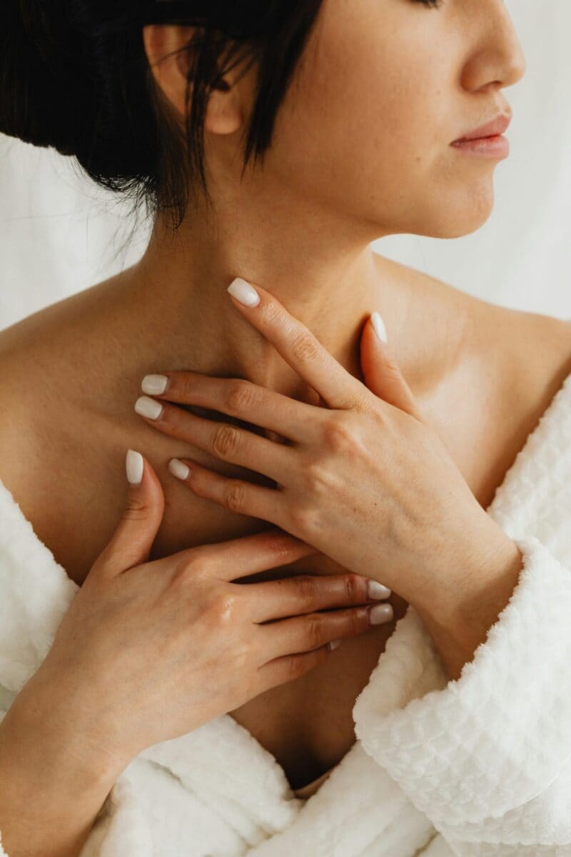 A woman in a white dressing gown touches her collarbone and neck with both hands, showing neatly manicured nails. Her face is partially visible, and she appears calm and relaxed—reflecting the confidence inspired by a specialist in plastic surgery.