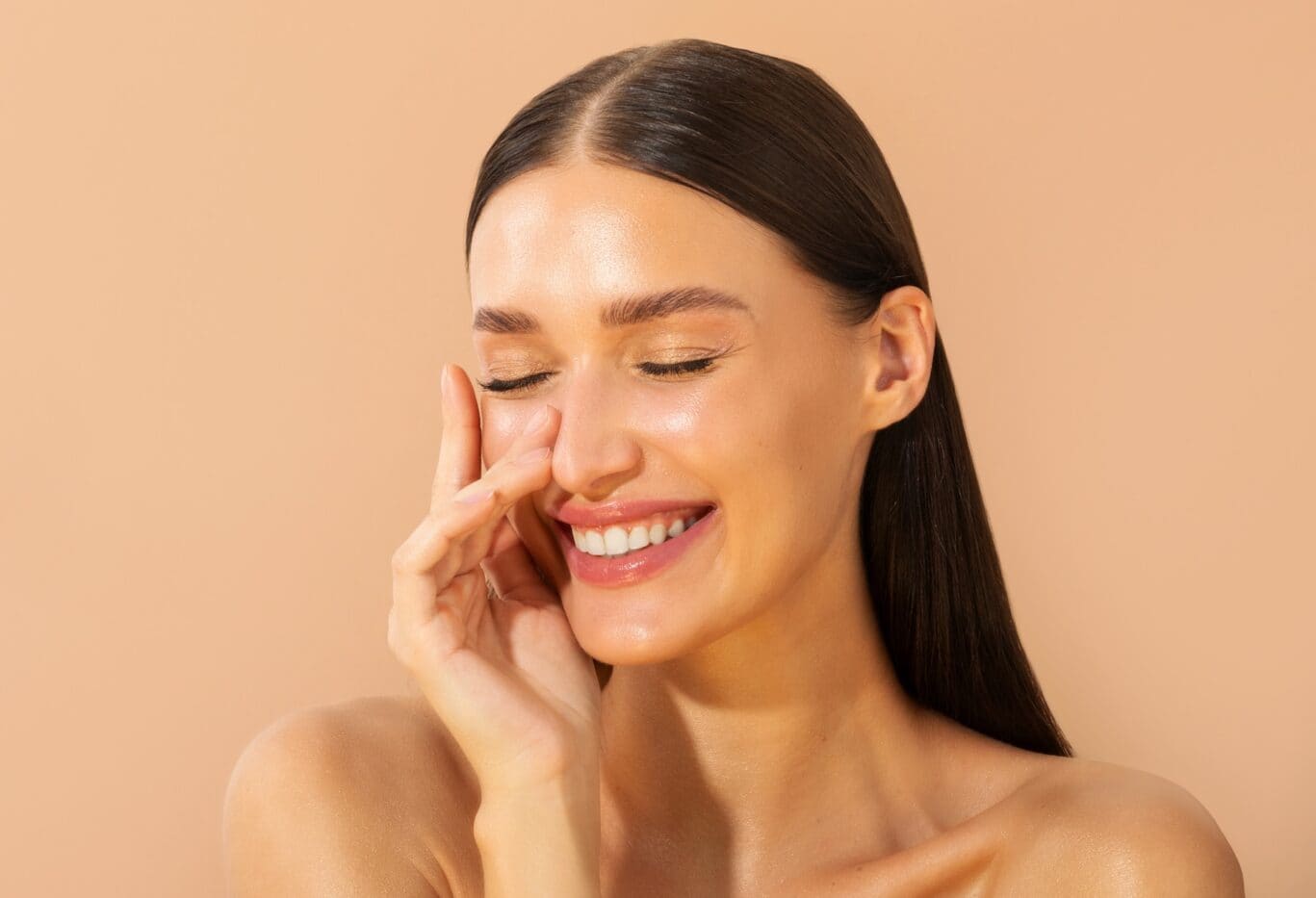 A woman with smooth, glowing skin and long brown hair smiles with her eyes closed, touching her face with one hand, against a beige background—radiating the confidence that comes from the care of a plastic surgery specialist.