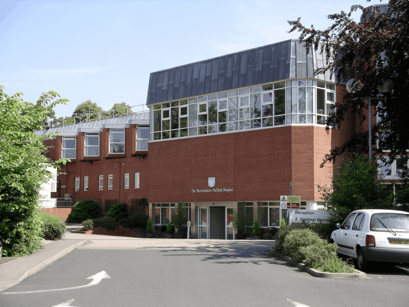 A modern red-brick hospital building with large windows, labelled The Newhall Hospital. Trees and greenery surround the entrance, and a hospital sign points towards reception, patient parking, and specialist clinics such as plastic surgery.