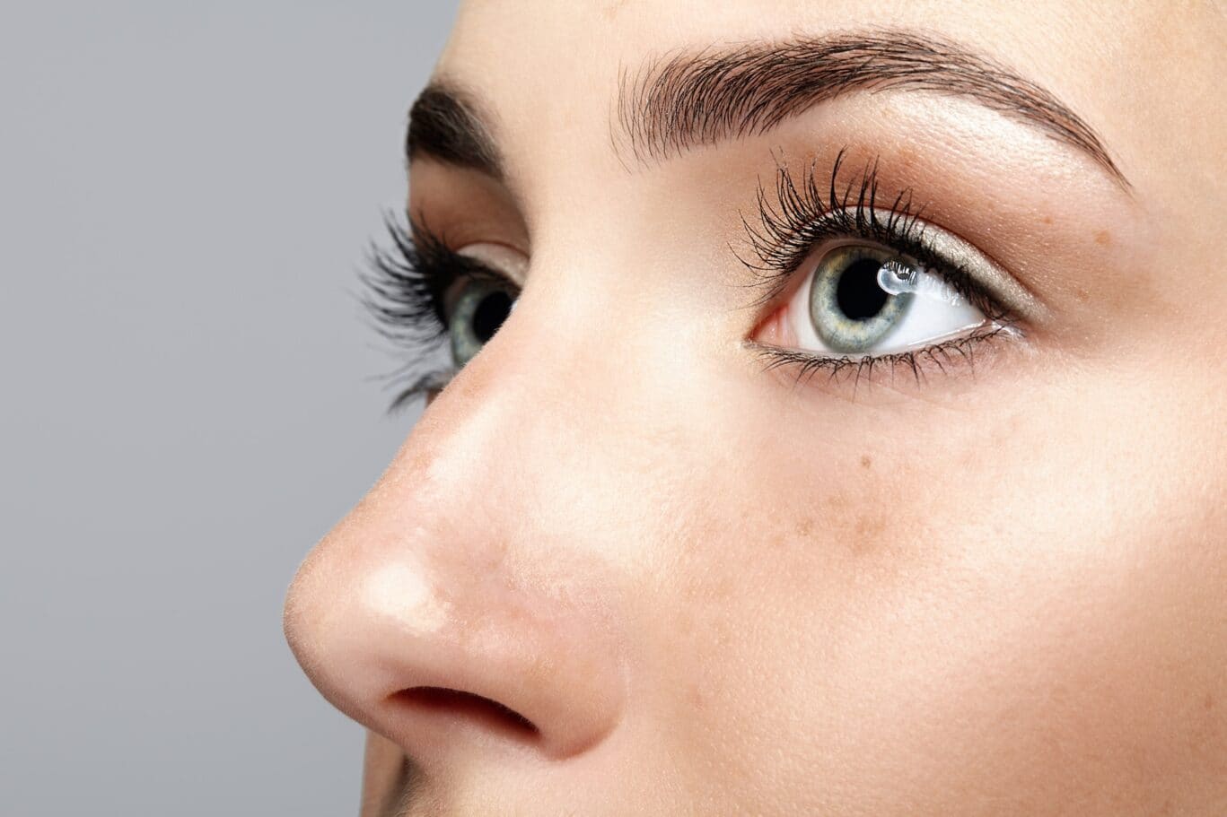 Close-up of a person's face focusing on blue-green eyes, long eyelashes, well-groomed eyebrows, and part of the nose against a plain light grey background, highlighting features that a plastic surgery specialist might emphasise during consultations.