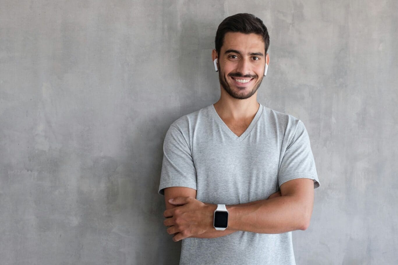Smiling man with short dark hair, wearing a grey t-shirt, white smartwatch, and wireless earbuds, stands with arms crossed against a plain grey textured wall, embodying the confidence of a plastic surgery specialist.