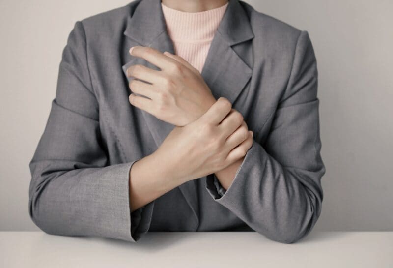 Person in a grey suit jacket and pink polo neck sitting at a white table, holding their right wrist with their left hand, suggesting discomfort or pain in the wrist—possibly awaiting consultation with a specialist. Face is not visible.