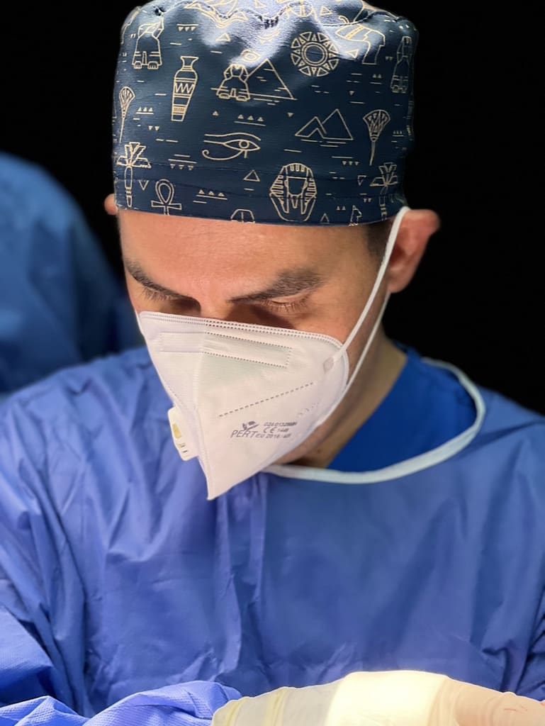 Consultant Plastic Surgeon Mr David Izadi wearing a face mask, blue scrubs, and a cap with Egyptian-themed designs prepares for a medical procedure.