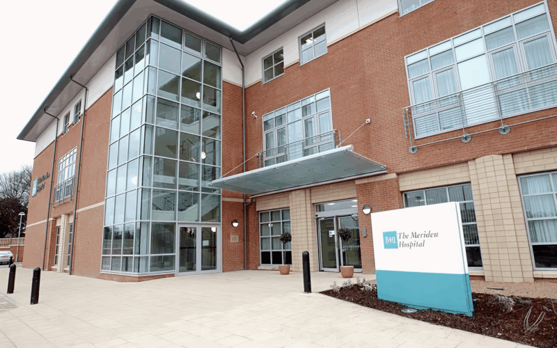 Modern hospital building with large windows, tan and red brick exterior, glass entrance canopy, and a sign reading The Meriden Hospital near the main doors. This facility is renowned for its specialist care in various fields, including plastic surgery.