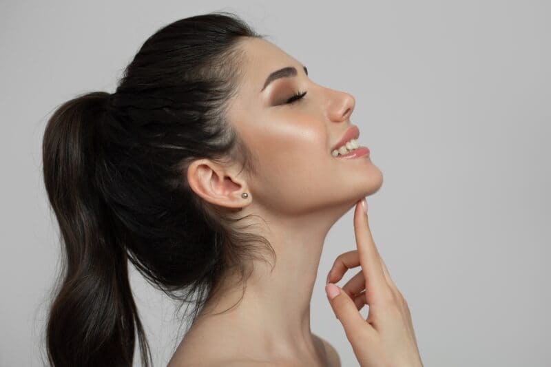 Woman with long dark hair in a ponytail smiling with eyes closed, tilting her head back and touching her chin with her finger against a plain light background, radiating confidence often seen after expert care from a plastic surgery specialist.