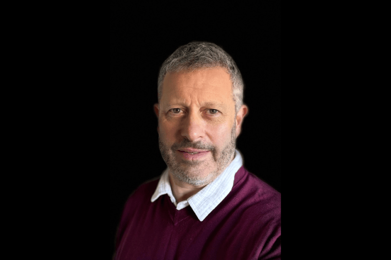 A middle-aged man with short grey hair and a beard, wearing a maroon jumper over a white collared shirt, poses against a black background, exuding the confident demeanour of a plastic surgery specialist.