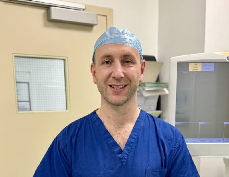 A plastic surgery specialist wearing blue medical scrubs and a disposable cap smiles in a hospital setting, standing in front of a door and medical equipment.
