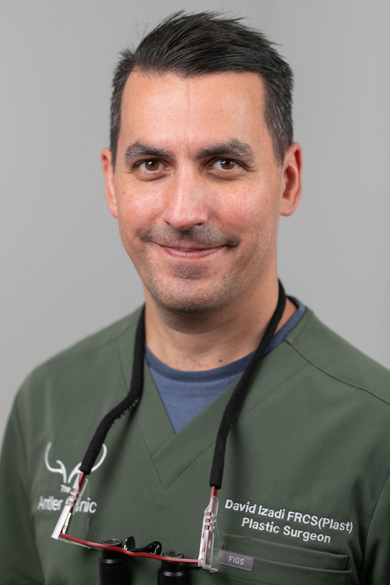 Consultant Plastic Surgeon Mr David Izadi wearing green medical scrubs and magnifying loupes around his neck, smiling at the camera. The name badge on his uniform indicates he is a plastic surgery specialist.