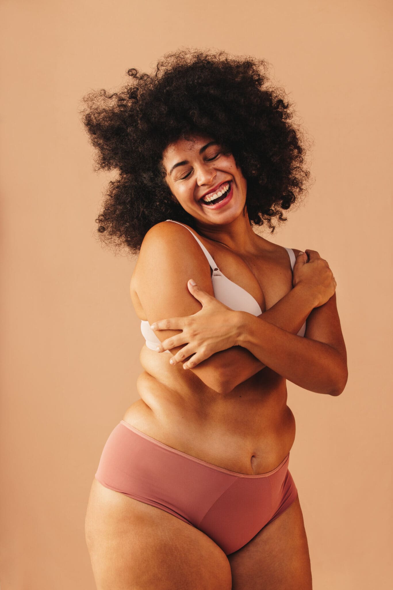 Woman with curly hair in a bra and knickers smiles joyfully, hugging herself against a plain peach background, radiating the confidence often seen after consulting a plastic surgery specialist.