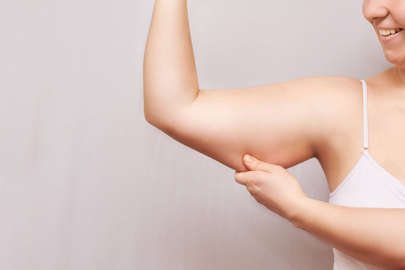 A person in a white vest flexes their arm and pinches the skin on their upper arm, showing loose skin or body fat. Smiling confidently, they seem to be considering options a specialist in plastic surgery might recommend, all against a plain background.