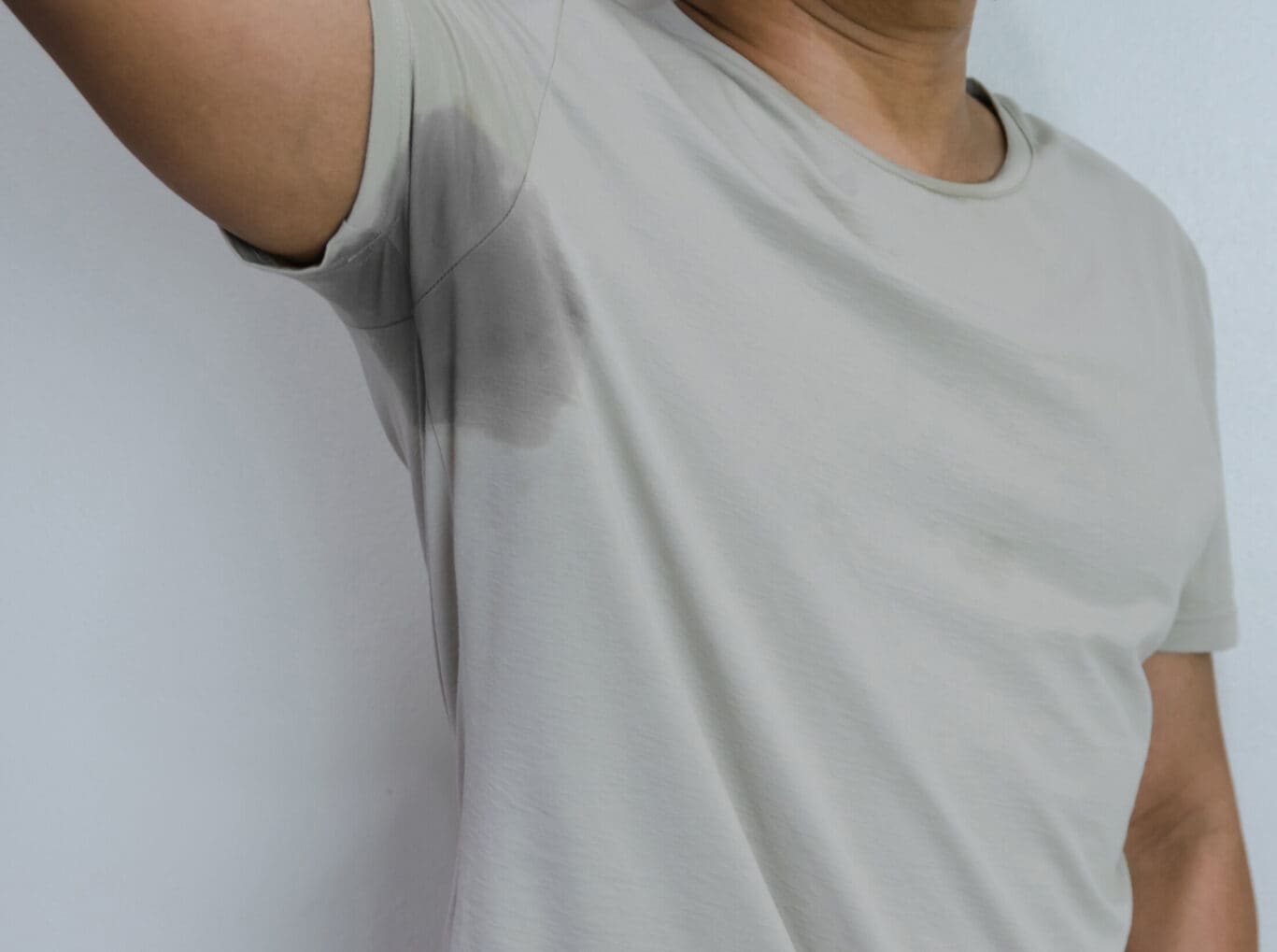 A person wearing a light grey t-shirt raises their arm, revealing a large sweat stain under the armpit. The background is plain and white, suggesting a scenario where consultation with a sweat management specialist or even plastic surgery might be considered.