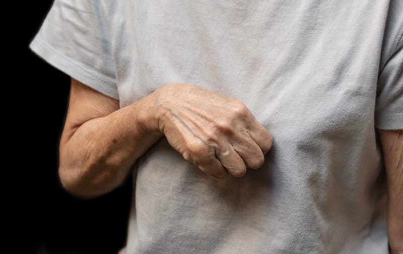 An older adult wearing a light grey t-shirt uses their right hand to make a fist with palm facing down, held in front of their chest against a dark background, as if demonstrating a gesture explained by a specialist.