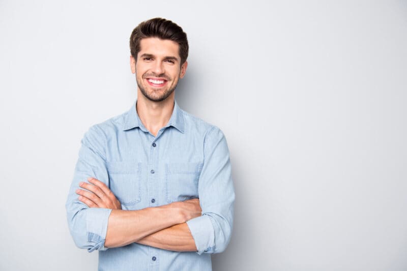 a man with short dark hair and a beard, wearing a light blue button-up shirt, stands smiling with his arms crossed against a plain light background.