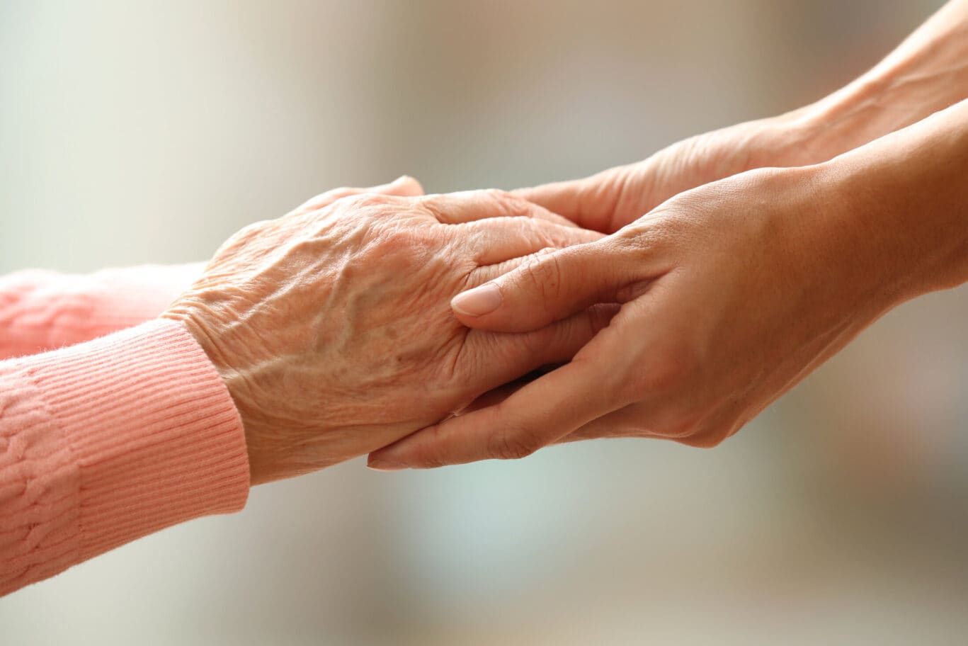 A younger person gently holds the hands of an older person wearing a pink jumper, conveying care, support, and connection—an image that beautifully reflects the compassionate bond often found between a specialist and their patient in the field of plastic surgery. The soft, blurred background enhances this heartfelt moment.