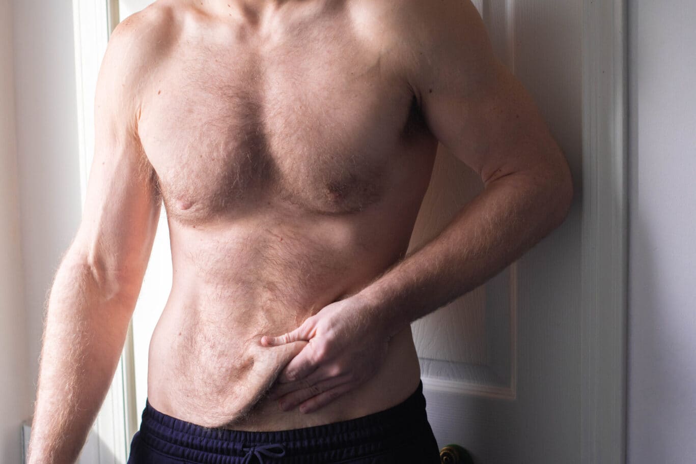 A shirtless person stands in front of a door, wearing dark shorts and pinching loose skin on their abdomen with one hand, perhaps considering options that a plastic surgery specialist might recommend.