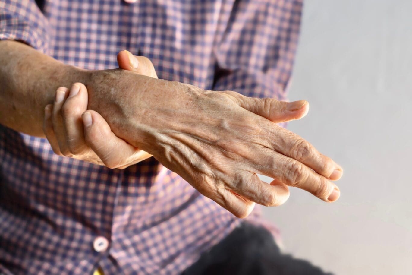 An elderly person wearing a checked shirt holds their painful wrist with the other hand, suggesting discomfort or joint pain, possibly arthritis—a condition that may require consultation with a specialist.