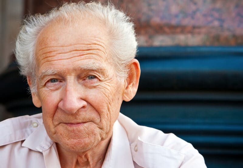 Elderly man with white hair wearing a light pink shirt, sitting outdoors and looking into the camera with a gentle expression, embodying the confidence often seen after consulting a plastic surgery consultant.