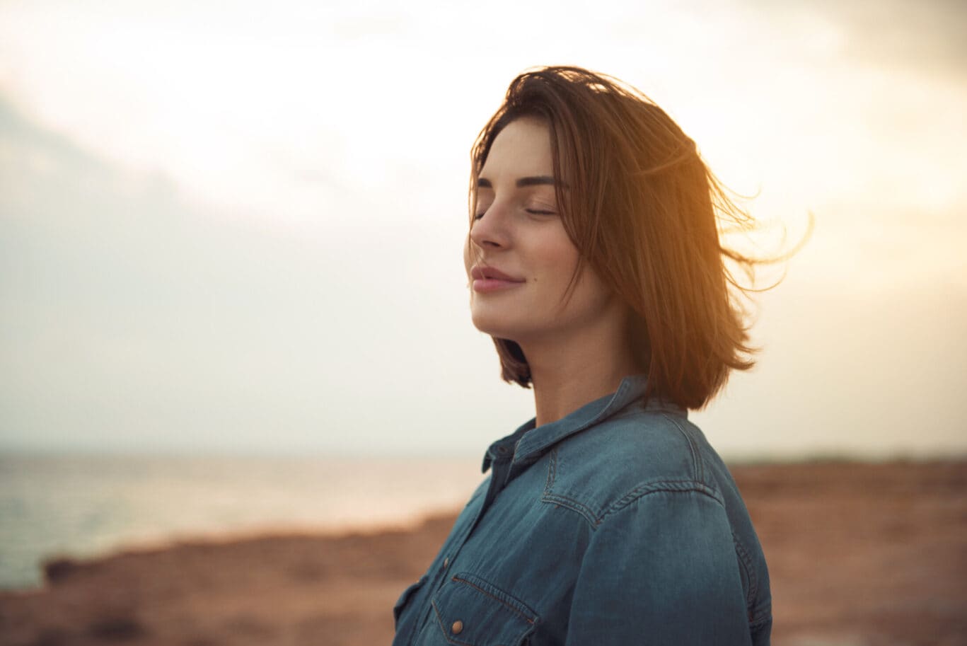 Charming calm young woman is standing near sea with closed eyes