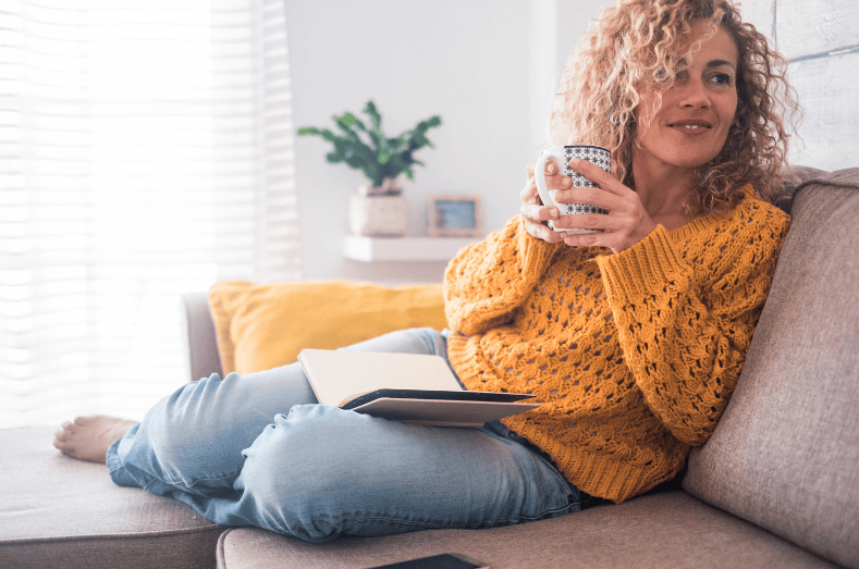 Woman with curly blonde hair in a yellow jumper sits on a sofa, holding a mug and smiling. An open notebook rests on her lap, perhaps filled with notes from her work as a plastic surgery specialist. Sunlight shines through a nearby window.